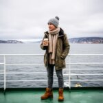 Cestovatel na trajektu do Švédska drží kávu, v pozadí moře a severské pobřeží za zatažené oblohy.
