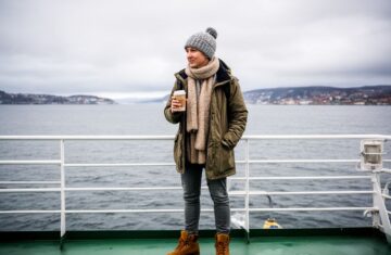 Cestovatel na trajektu do Švédska drží kávu, v pozadí moře a severské pobřeží za zatažené oblohy.