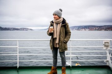 Cestovatel na trajektu do Švédska drží kávu, v pozadí moře a severské pobřeží za zatažené oblohy.